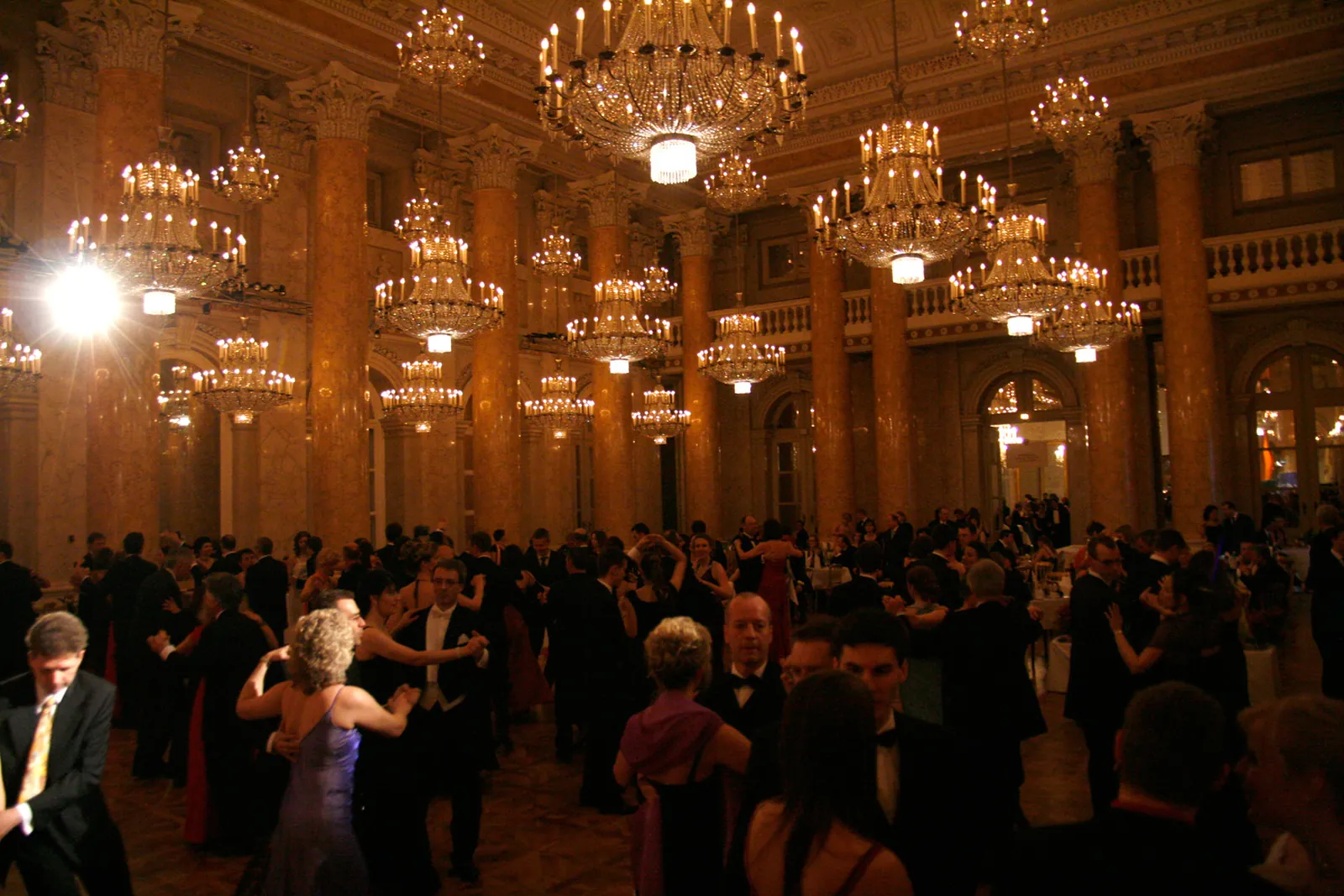 Der Zeremoniensaal der Hofburg während eines Balls, mit hohen Marmorsäulen, zahlreichen Leuchtern und tanzenden Paaren