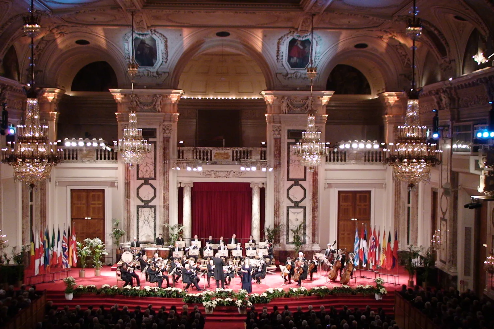 Der Hofburg Festival Hall von der Galerie aus, mit Kristallleuchtern, roten Marmorpilastern, einem purpurroten Vorhang und dem Orchester auf einer rot ausgelegten Bühne