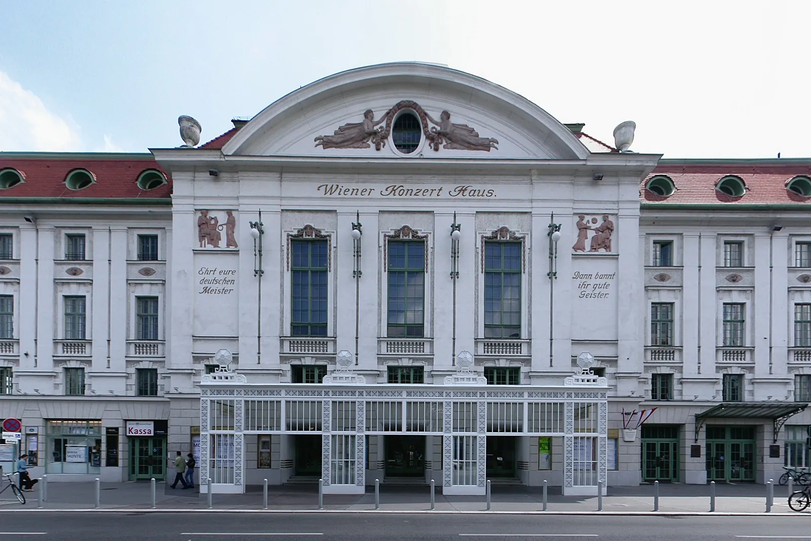 Die weiße Straßenfassade des Wiener Konzerthauses, mit rotem Ziegeldach und einer Glas-Eisen-Vorhalle über den Eingängen