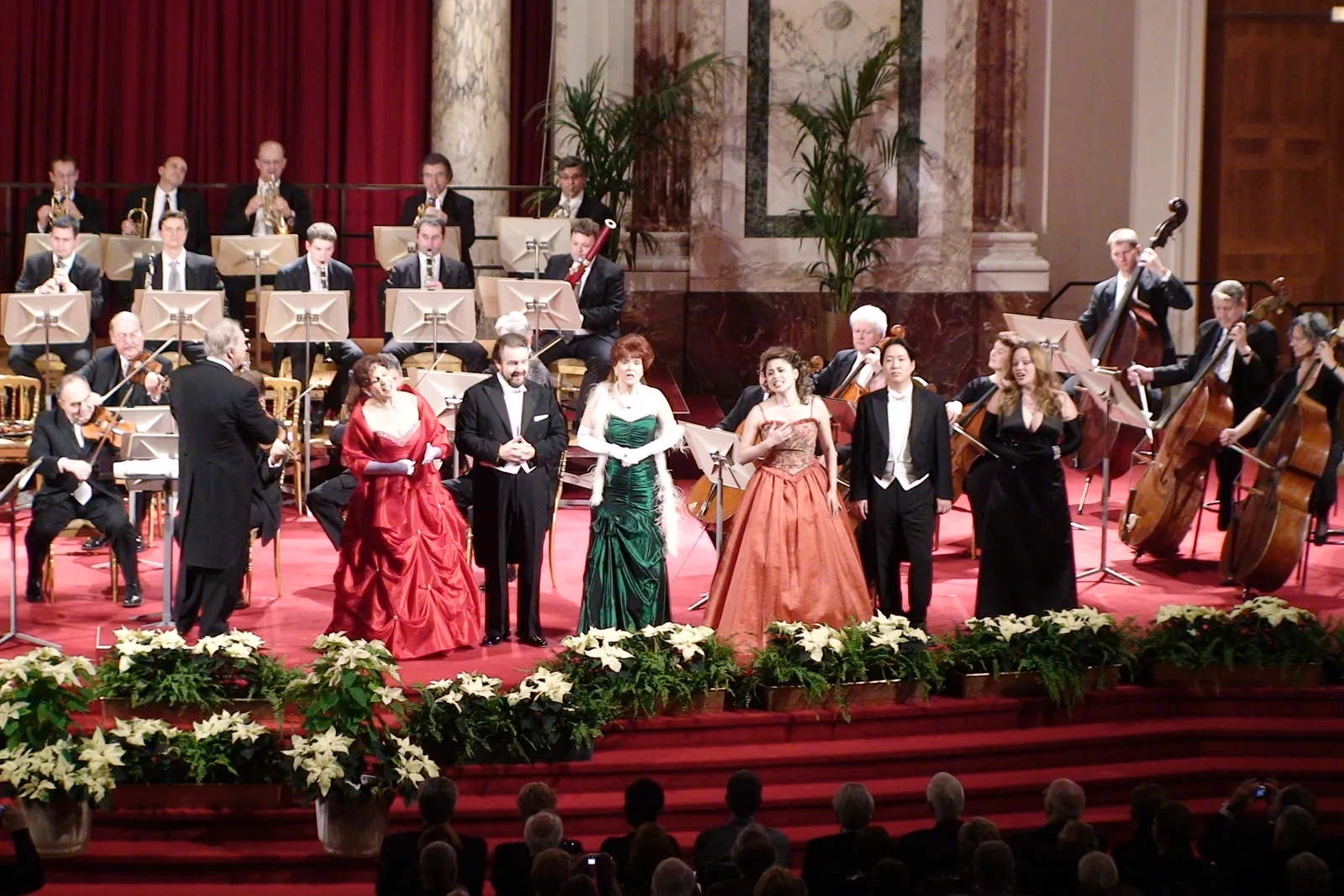 Sechs Opernsolisten in roten, grünen, orangefarbenen und schwarzen Roben und im Abendanzug stehen vor den Streichern auf der Bühne des Hofburg Festival Hall