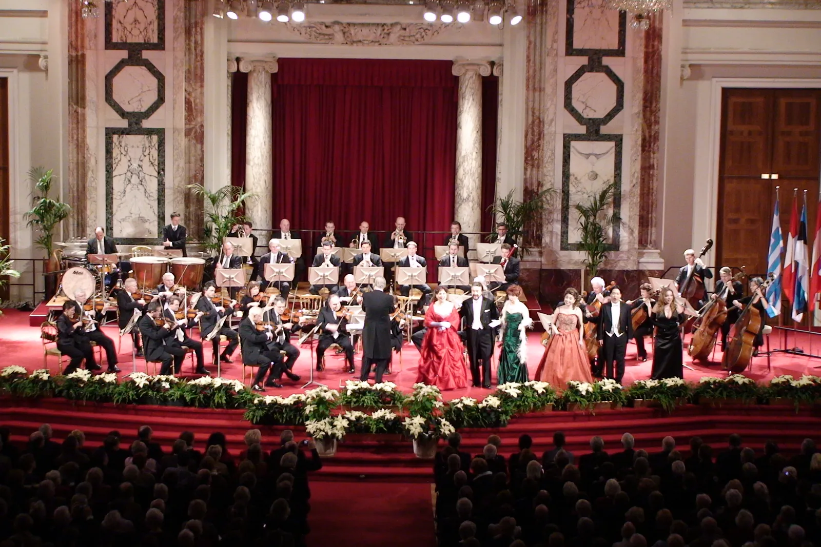 Das Vienna Hofburg Orchestra und fünf Solisten im Abendanzug auf einer rot ausgelegten Bühne im Hofburg Festival Hall, mit purpurrotem Vorhang und Marmorpilastern im Hintergrund