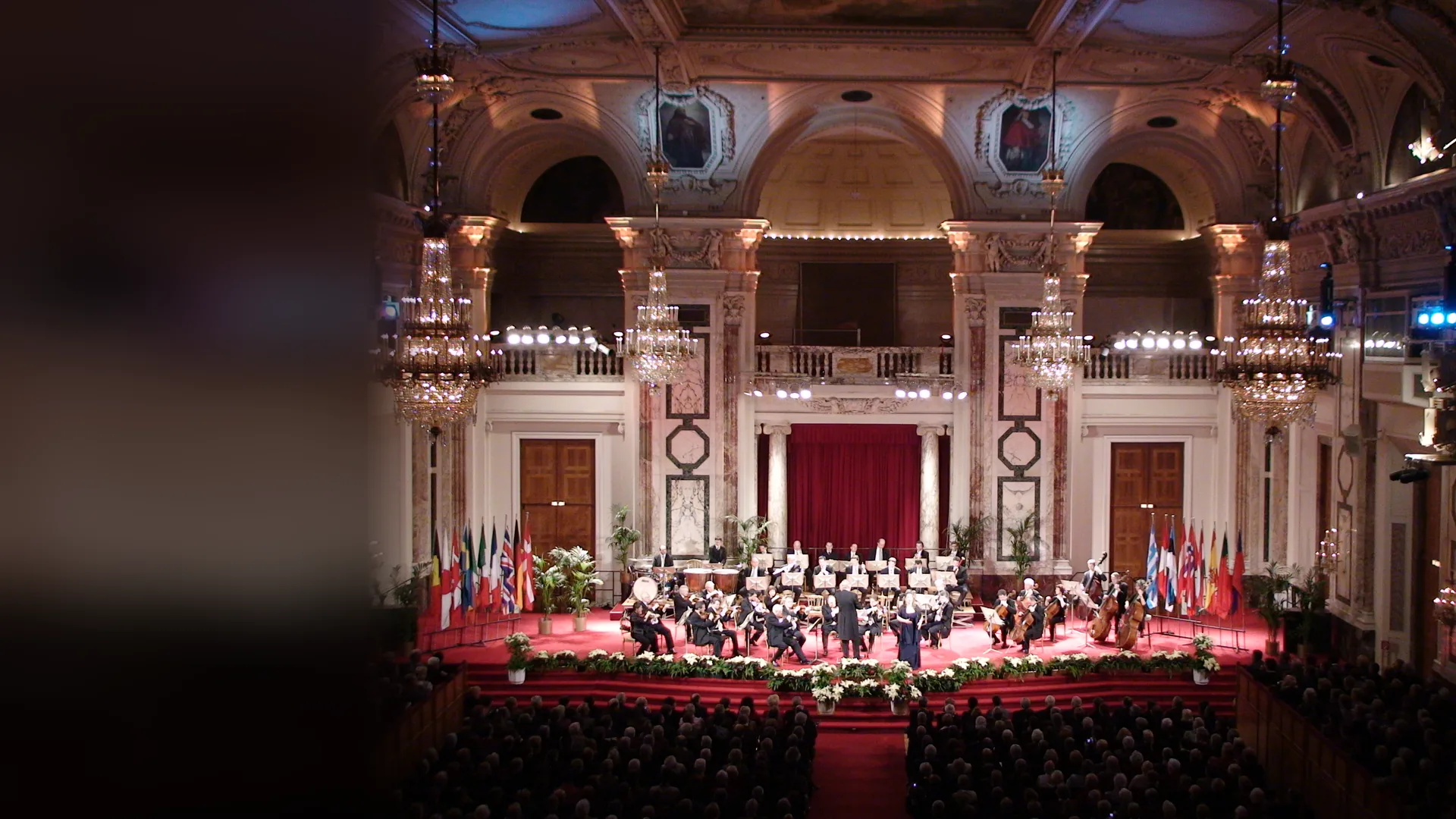 Vídeňský hofburgský orchestr na červeném koberci pódia ve slavnostním sále Hofburgu, pod křišťálovými lustry, mramorovými pilastry a karmínovým jevištním závěsem, s publikem vpředu